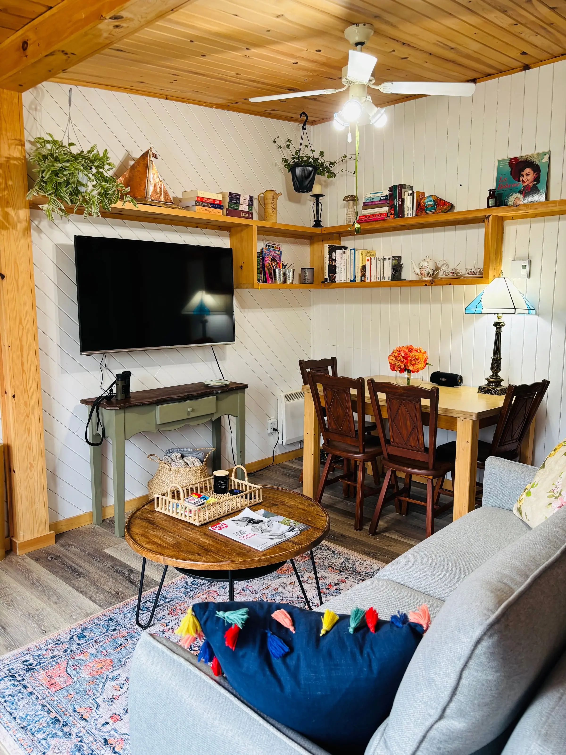Le salon du chalet Encore un peu... : canapé gris, table basse ronde, télévision, étagères de pin garnies de livres et coin repas