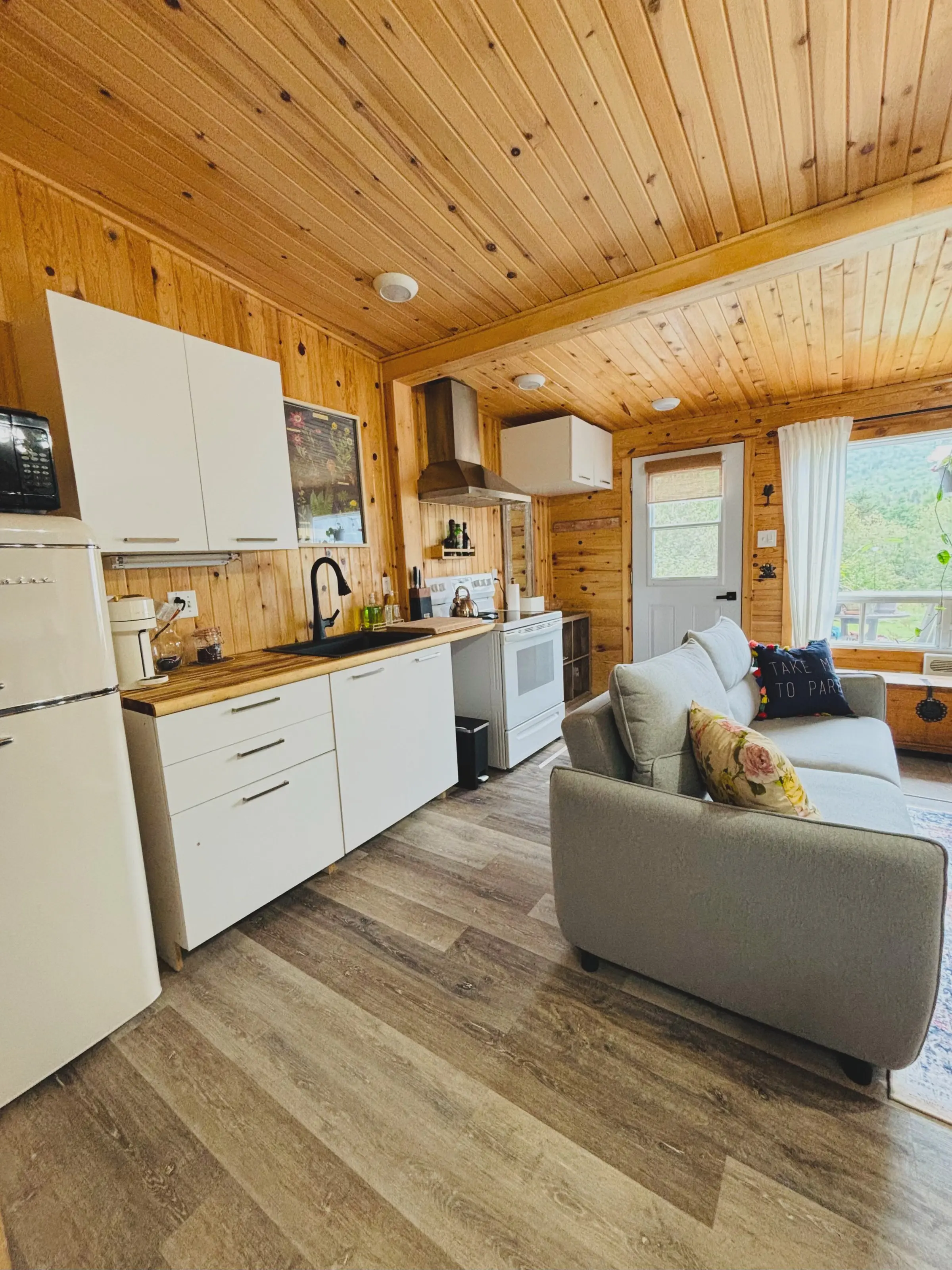 La cuisine blanche au comptoir de bois et le canapé gris du chalet Encore un peu..., sous le plafond de pin, la fenêtre ouverte sur la montagne